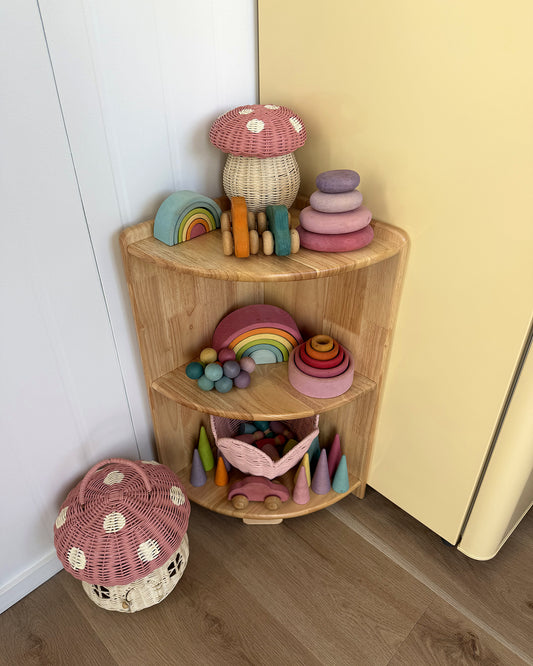 Angled view of the JORA Corner 3 three-tier wooden toy shelf in a playroom corner, styled with wooden toys