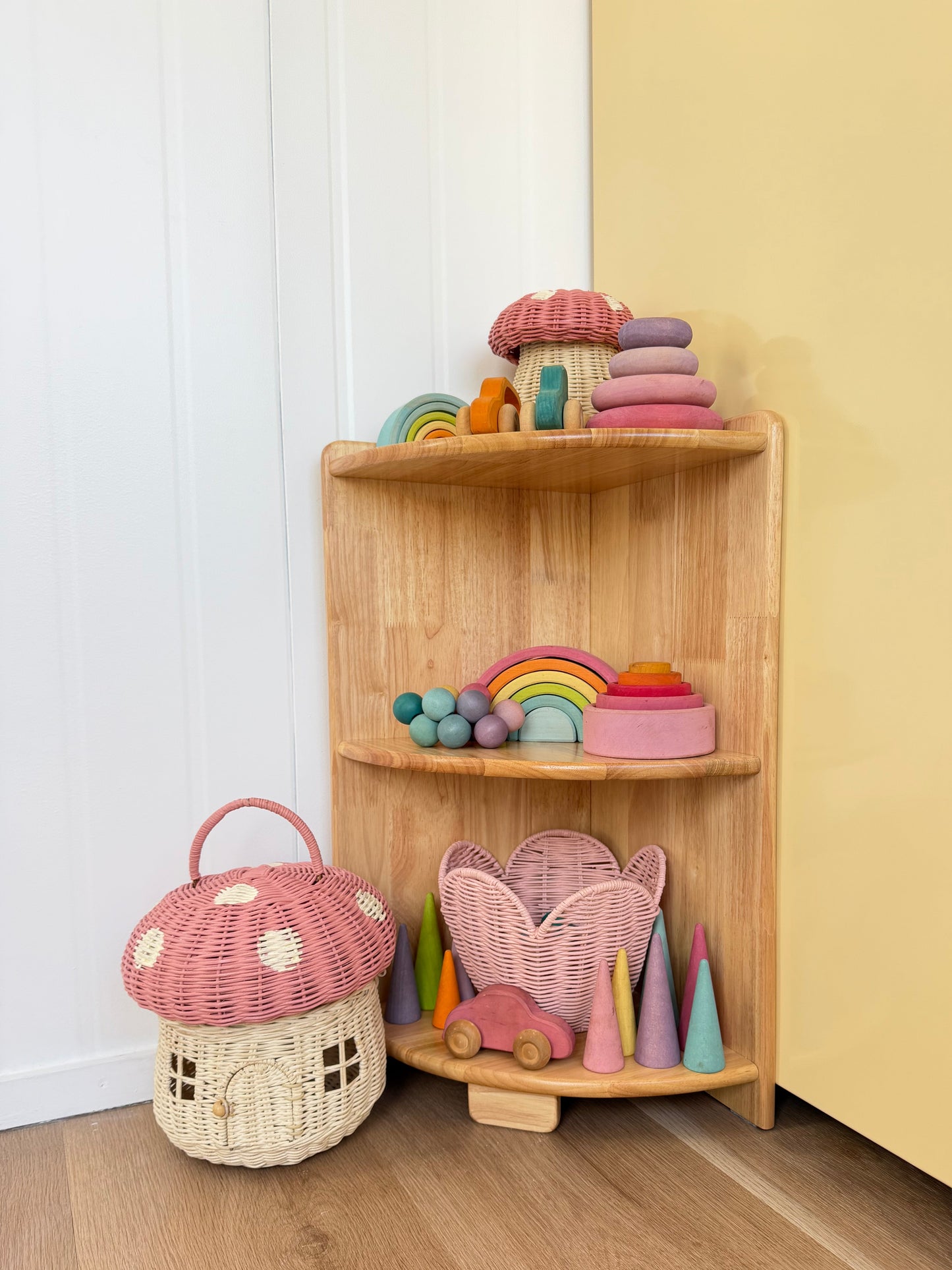 A corner wooden toy shelf styled in a nursery or playroom corner, holding colorful wooden toys including a rainbow, cones, a mushroom house