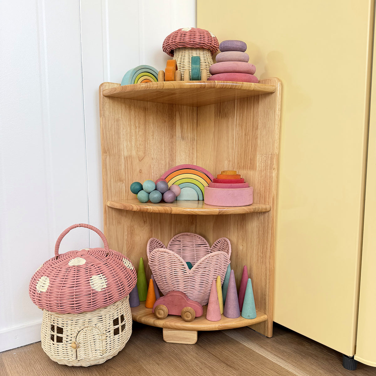 A corner wooden toy shelf styled in a nursery or playroom corner, holding colorful wooden toys including a rainbow, cones, a mushroom house