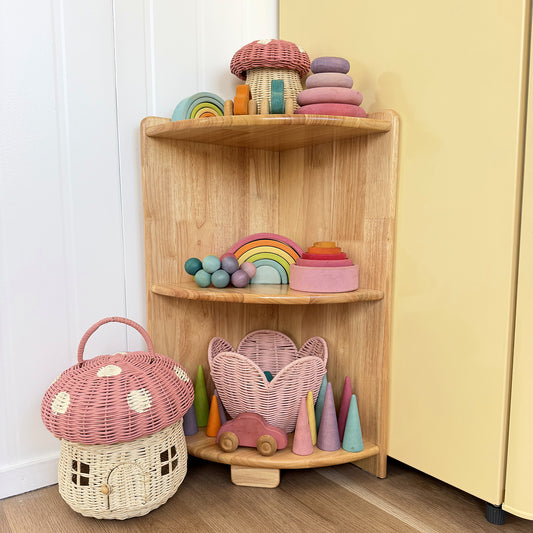 A corner wooden toy shelf styled in a nursery or playroom corner, holding colorful wooden toys including a rainbow, cones, a mushroom house