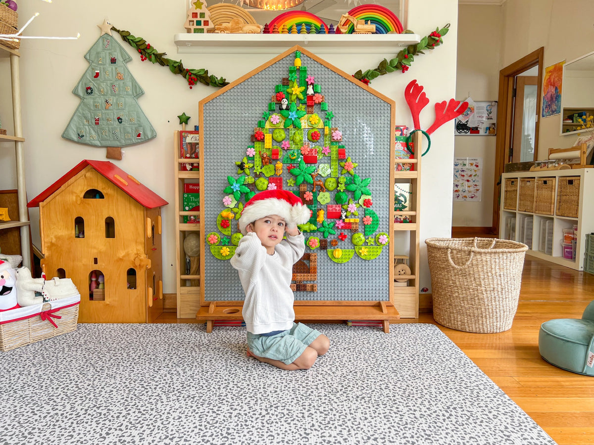 The house-shaped Building Board standing free in a playroom, a Christmas tree built on its baseplate face from DUPLO bricks, a toddler sitting in front of it