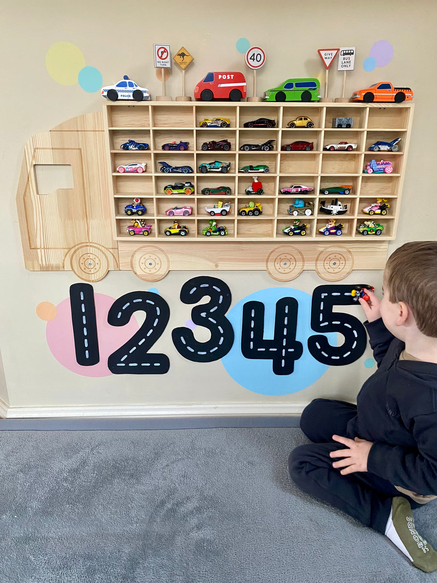 The JCAR Truck rack mounted on a playroom wall, filled with 1:64 cars, a child sitting below with road-shaped number cutouts