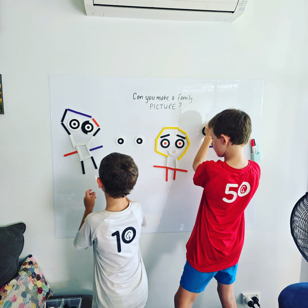 Two children drawing faces in marker on a white JoyKids magnetic decal, with the question "can you make a family picture?" written above